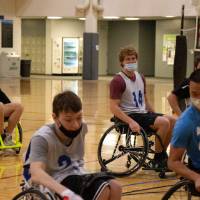 Adult dribbling a basketball in a wheelchair with other participants defending the ball.